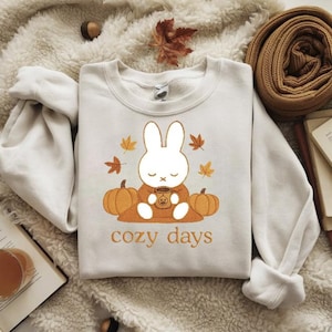 Op de afbeelding: Crèmekleurige sweatshirt met een cartoonkonijn dat een mok vasthoudt, omringd door pompoenen en herfstbladeren. De tekst "cozy days" staat eronder in oranje. Een herfstthema.