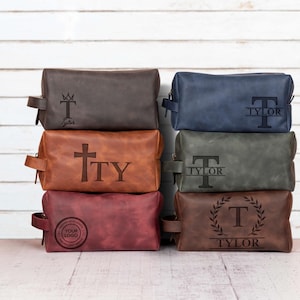 May include: A stack of six leather toiletry bags in various colors, including brown, blue, and burgundy. Each bag features a unique engraved design, such as initials, a cross, or a logo. The bags have a rectangular shape with a handle on the side.