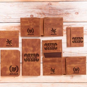 May include: A collection of brown leather wallets and cardholders. Some items feature the name "JULIAN" and various designs, including a leaf and the outline of Ohio. The items are arranged on a light wood surface.
