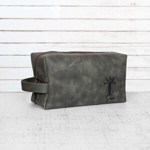 May include: A gray leather toiletry bag with a zipper closure and a handle. The bag has a black graphic of a crown and the letter "T" with the name "Tyler" below it.
