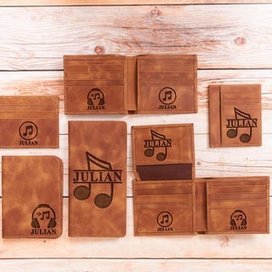 May include: A collection of brown leather wallets, cardholders, and a notebook. Each item is engraved with the name "JULIAN" and musical symbols, including headphones and musical notes. The items are arranged on a light wood surface.