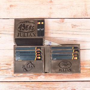 May include: Gray leather wallet with multiple card slots and a bill compartment. The wallet is personalized with the name "JULIAN" and the "49ERS" logo and football graphic. The wallet is open, revealing credit cards and cash.