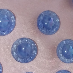 May include: Close-up of several light blue, round glass beads. Each bead has a textured, iridescent center and a ring of small, clear, bubble-like details around the edge. The beads are arranged on a soft, pink fabric.