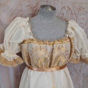 May include: A cream-colored dress featuring short puffed sleeves with gold trim, a square neckline, and a bodice adorned with a metallic gold floral pattern. A solid gold ribbon encircles the waist, separating the bodice from the plain skirt.