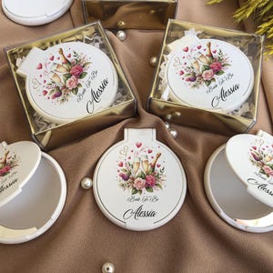 May include: White compact mirrors with a floral and champagne bottle design, labeled "Bride To Be Alessia." Some mirrors are in clear boxes with gold trim. The design includes pink and red flowers and hearts.