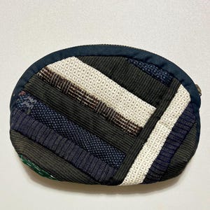 以下が含まれることがあります： パッチワークデザインの小さな楕円形の小銭入れ。ネイビー、黒、白、茶色の様々な生地が対角線ストライプに配置されています。ネイビーブルーのジッパーとトリムがデザインを完成させています。