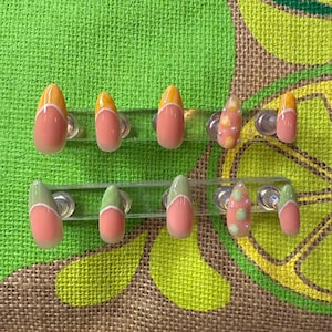 May include: A set of ten artificial nails in various designs. Some nails feature a peach base with yellow or green tips, while others have floral or polka dot accents. The nails are displayed on clear plastic holders against a green textured background.