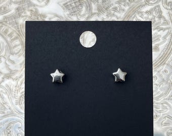 Op de afbeelding: Een paar kleine, gepolijste, zilverkleurige oorknopjes in de vorm van een ster. De oorbellen zijn bevestigd op een rechthoekige zwarte displaykaart met een rond gat in het midden aan de bovenkant om ze op te hangen.