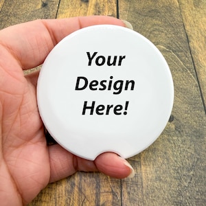 May include: A white, round car coaster with a curved notch for easy removal. The text "Your Design Here!" is printed in black, centered on the coaster. The coaster is held in a hand against a wooden background.