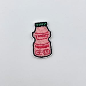 Op de afbeelding: Geborduurde patch in de vorm van een roze yoghurtfles met een groene dop. Het woord "Yogurt" is rood geborduurd op de voorkant. De patch heeft een zwarte rand en tekst in een vreemde taal.