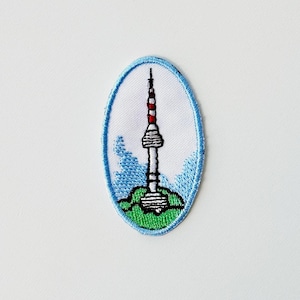 May include: Embroidered patch featuring a stylized depiction of a tower with a red and white top, set against a blue sky and green ground. The patch has a blue border and is oval-shaped.