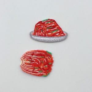 以下が含まれることがあります： 鮮やかな赤と緑のキムチのデザインが施された刺繍パッチ。1つのパッチは銀色の皿に盛られたキムチの山、もう1つはキムチの葉1枚を描いています。衣類やアクセサリーを個性的に彩るのに最適です。