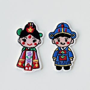 May include: Two embroidered patches featuring cartoon figures in traditional Korean attire. The female figure wears a red and purple dress with a green headdress. The male figure is in a blue and gold outfit with a matching hat. Both have rosy cheeks.