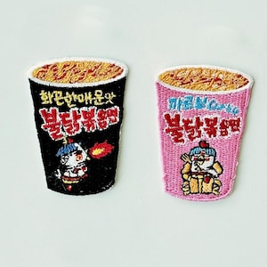Puede incluir: Dos parches bordados con forma de tazas de ramen instantáneo. Uno es negro, el otro rosa. Ambos tienen texto coreano y un personaje de dibujos animados. Para ropa.