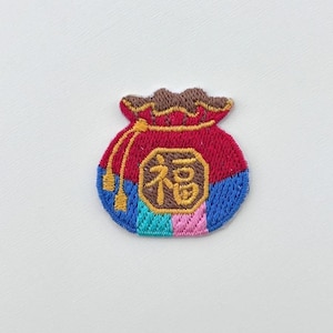 May include: Embroidered patch of a pouch. The pouch is red with a brown top and a blue and multi-coloured bottom. A gold emblem with Asian characters is centered on the pouch. Gold tassels hang from the top. Ideal for clothing or accessories.