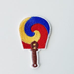 May include: Embroidered patch featuring a stylized fan design. The fan has a red, yellow, and blue spiral pattern with a brown handle. The patch is outlined in white and is likely for clothing or accessories.