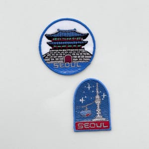 May include: Two embroidered patches featuring Seoul landmarks. One is a circular patch with a blue and white design of a traditional building. The other is an arched patch with a blue background, a tower, and the word "SEOUL" in red.