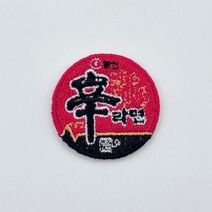 May include: Round embroidered patch with a red background and black and white text. The patch features Asian characters and the word "ramen". The patch has a black and white border.