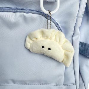 May include: A plush, cream-colored dumpling charm with black dot eyes hangs from a silver chain and white plastic clip. The dumpling is attached to a light blue backpack with a zipper and pocket.