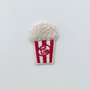 Puede incluir: Parche bordado de palomitas de maíz. Presenta un recipiente a rayas rojas y blancas con las palabras "Pop Corn" en un óvalo blanco. Las palomitas son de color blanco roto y esponjosas.