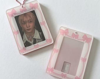May include: Two rectangular photo card holders. One contains a photo of a person with light-colored hair and a necklace. Both holders are clear with a pink bow design around the edges and a pink keychain.