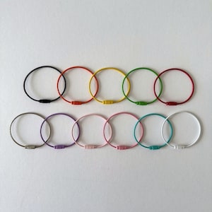 May include: Ten colourful key rings are displayed against a white backdrop. The rings are in a variety of colours, including black, red, yellow, and green. Each ring has a circular design with a metal clasp. The key rings are arranged in two rows.