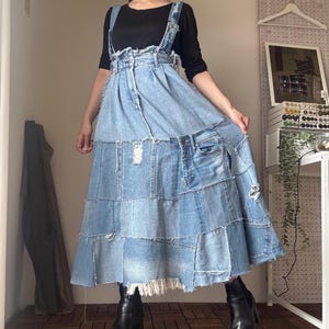 以下が含まれることがあります： 明るいブルーデニムのオーバーオールワンピース。ティアードスカートが特徴です。パッチワークデザインで、縫い目やダメージ加工が施されています。トップスは黒の長袖シャツ。黒いブーツを履いています。