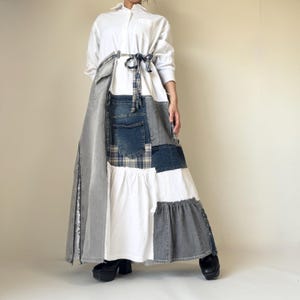 Op de afbeelding: Een patchwork denim en witte jurk met een witte overhemdtop met kraag. De rok heeft verschillende denim patches in blauwe en grijze tinten, met een witte gerimpelde sectie. Een blauw-witte geruite stropdas sluit de taille af.