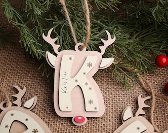 May include: A wooden Christmas ornament shaped like a reindeer with the letter "K" cut out. The ornament is light pink with white accents and the name "Kristin" written on it. It hangs from a twine string.