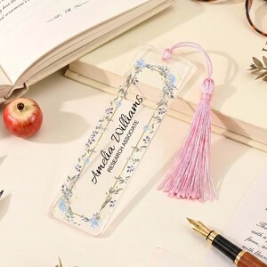 May include: A clear rectangular acrylic bookmark featuring a printed floral wreath border in shades of blue and green. The text Amelia Williams and Research Associate is printed in black. A light pink silk tassel is attached to the top.