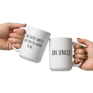 May include: Two white ceramic coffee mugs. One mug reads "THIS COFFEE SMELLS LIKE YOU'RE GOING TO BE..." and the other reads "AN UNCLE!". The mugs are held up in a celebratory gesture.