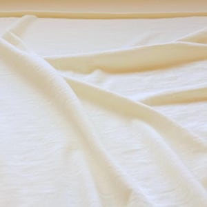 May include: A solid off-white fabric with a soft, slightly textured surface. The material is draped in loose, flowing folds, creating subtle shadows and highlights across the surface. The textile appears lightweight and opaque with a matte finish.