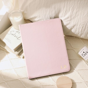 May include: A pale pink tablet case with gold initials "L.B." on a white knitted blanket. A wooden coaster, a candle, and a book are also visible. The case has a stitched border.