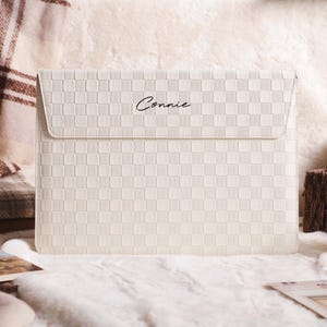 May include: A cream-colored laptop sleeve with a checkered pattern and a flap closure. The word "Connie" is written in cursive on the front. The sleeve is made of a textured material and is designed to protect a laptop.