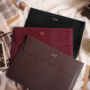 May include: Three personalised leather laptop sleeves in black, burgundy, and brown, each with embossed initials. The sleeves have a crocodile skin texture. A cup of coffee and a postcard are in the background.