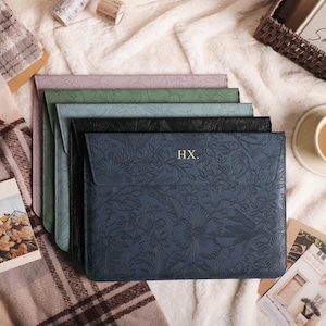 May include: Five laptop sleeves in a stack, in shades of lavender, green, blue, black, and navy. The navy sleeve has a floral design and the initials "HX." A cup of coffee, postcards, and a woven basket are also visible.