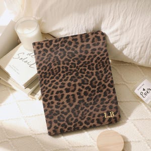 Può includere: Un diario con stampa leopardata marrone e nera con le iniziali dorate "L.B." è esposto su una superficie color crema. Sono visibili anche un piccolo coperchio di legno e una candela in un barattolo di vetro. Il diario è un accessorio elegante.