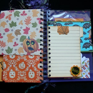May include: Open spiral-bound notebook with Halloween-themed pages. One page features a pumpkin illustration and autumn leaves. Another page has lined paper with a spiderweb-shaped paperclip. Additional pages have Halloween patterns and the word "Boo!"