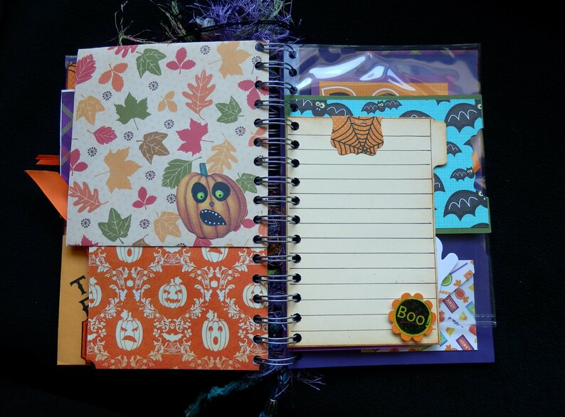 May include: Open spiral-bound notebook with Halloween-themed pages. One page features a pumpkin illustration and autumn leaves. Another page has lined paper with a spiderweb-shaped paperclip. Additional pages have Halloween patterns and the word "Boo!"