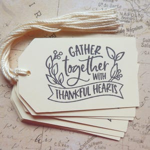 May include: A stack of cream-coloured rectangular paper tags with clipped corners, featuring a dark grey printed design of leaves and the text GATHER together WITH THANKFUL HEARTS. Each tag has a white string loop attached at the top.