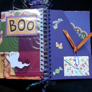 May include: Open spiral-bound Halloween scrapbook with decorative pages. Features include stickers, pockets, and the word "BOO" on a card. Other decorations include a ghost, candy corn, and a "Trick or Treat" ribbon.