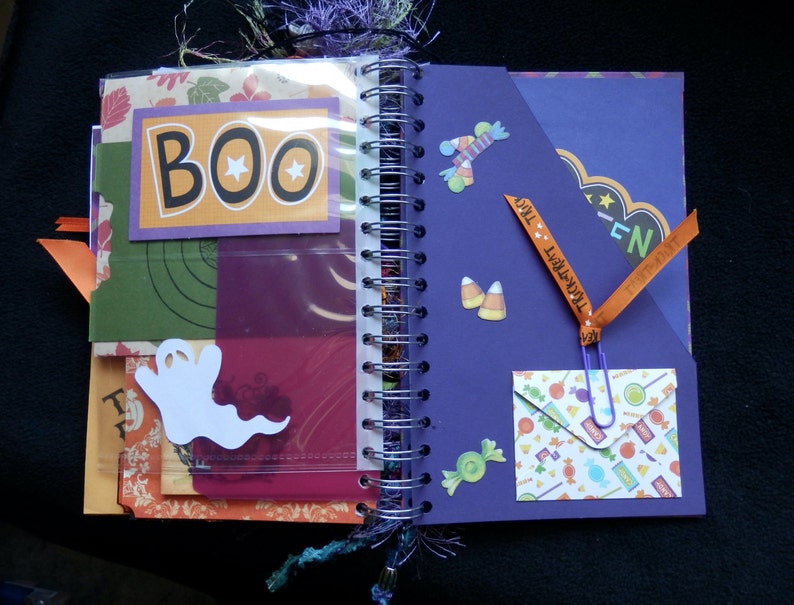 May include: Open spiral-bound Halloween scrapbook with decorative pages. Features include stickers, pockets, and the word "BOO" on a card. Other decorations include a ghost, candy corn, and a "Trick or Treat" ribbon.