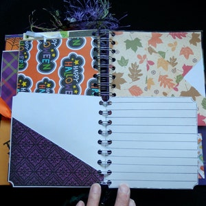 May include: Open spiral-bound journal with lined pages and decorative inserts. The inserts feature Halloween and autumn-themed patterns in orange, purple, and leaf designs. An orange ribbon and purple tassel add accents. The journal is open, revealing the lined pages.