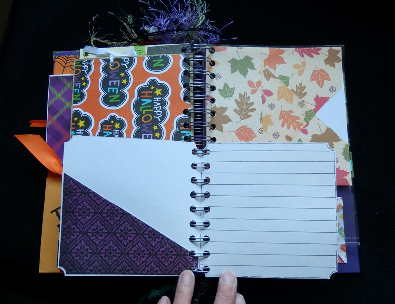 May include: Open spiral-bound journal with lined pages and decorative inserts. The inserts feature Halloween and autumn-themed patterns in orange, purple, and leaf designs. An orange ribbon and purple tassel add accents. The journal is open, revealing the lined pages.