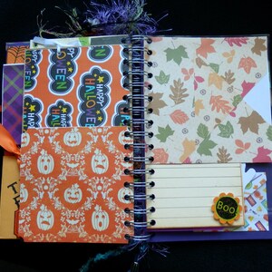 May include: Open spiral-bound journal with Halloween-themed pages. Features orange and white pumpkin patterns, leaf designs, and "Happy Halloween" text. Includes an orange ribbon and a "Boo!" embellishment.