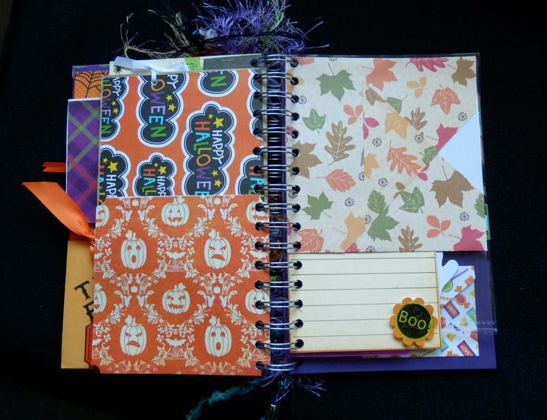 May include: Open spiral-bound journal with Halloween-themed pages. Features orange and white pumpkin patterns, leaf designs, and "Happy Halloween" text. Includes an orange ribbon and a "Boo!" embellishment.