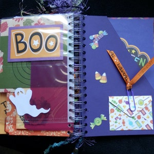 May include: Open Halloween scrapbook with a spiral binding. The pages feature colourful paper with decorative elements like a white ghost, candy corn stickers, and a "BOO" sign. Orange ribbon with "Trick or Treat" text is attached.