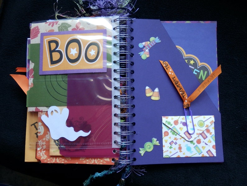 May include: Open Halloween scrapbook with a spiral binding. The pages feature colourful paper with decorative elements like a white ghost, candy corn stickers, and a "BOO" sign. Orange ribbon with "Trick or Treat" text is attached.