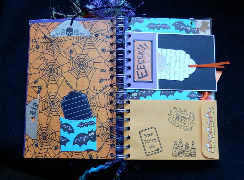 May include: Halloween scrapbook open to a double-page spread. One page has an orange spiderweb design, the other a blue bat pattern. Includes tags, pockets, and the text "EEEK!!" and "Witches Brew". Features a "Broom Parking Only" sign.