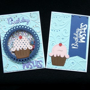 May include: Two handmade birthday cards with a light blue background and cupcake designs. One card features a shaker element with confetti. The cards have the words "Birthday Wishes" in a script font.
